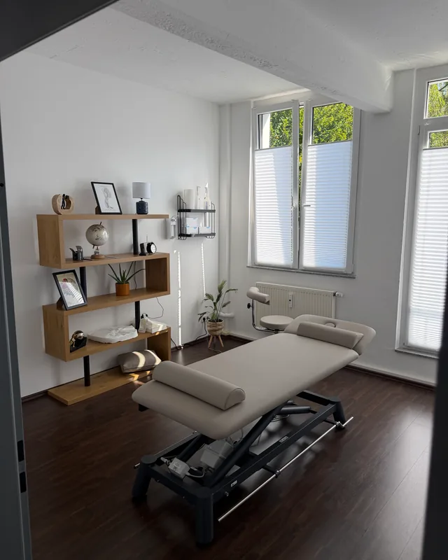 Behandlungsraum mit Liege, Holzregal und zwei Fenstern
